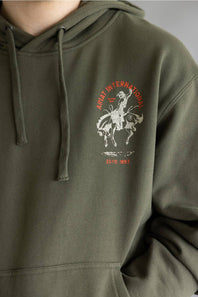 Close-up of an olive green hoodie with a drawstring hood and a chest graphic featuring a cowboy on a bucking horse in white with red text reading 'ARAT INTERNATIONAL' and 'ESTD. 1993'.