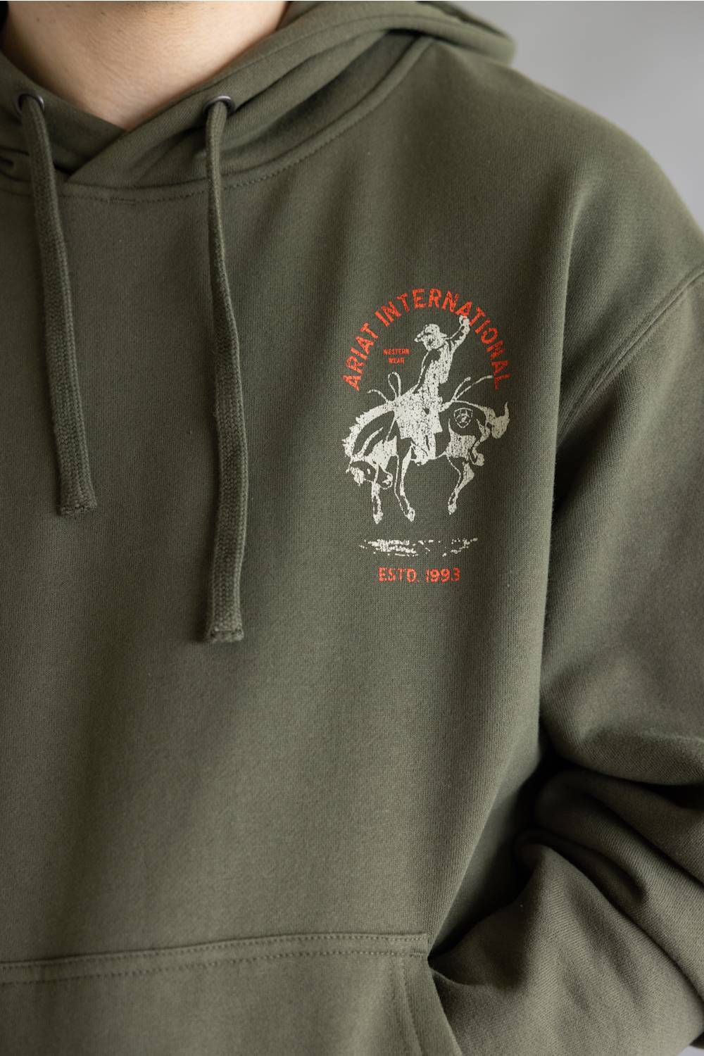 Close-up of an olive green hoodie with a drawstring hood and a chest graphic featuring a cowboy on a bucking horse in white with red text reading 'ARAT INTERNATIONAL' and 'ESTD. 1993'.