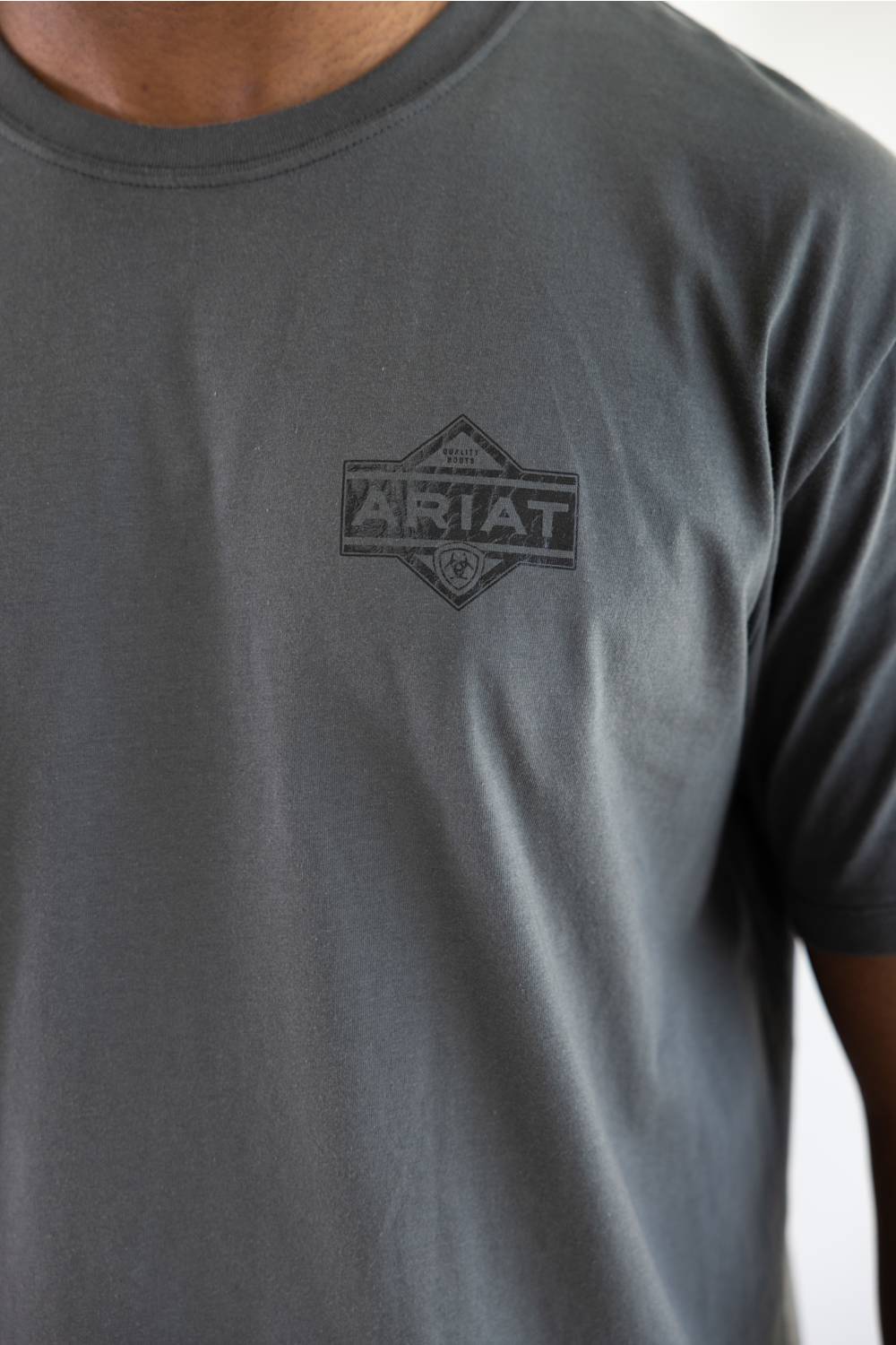 A close-up of a person wearing a dark gray crewneck t-shirt with a tonal dark gray 'Ariat' logo printed on the left chest.