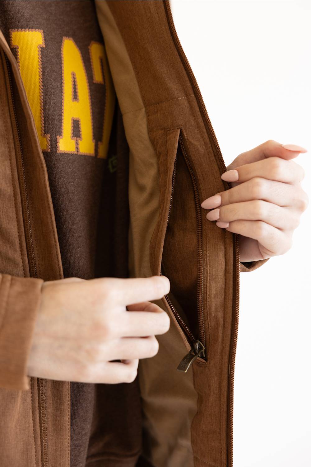 Close-up of a person wearing a brown zip jacket over a brown sweatshirt with yellow lettering, showing the interior pocket and zipper detail.