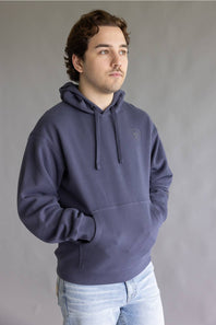 A man wearing a slate blue pullover hoodie with a small tonal embroidered logo on the left chest, paired with light-wash jeans, standing against a neutral gray studio background.