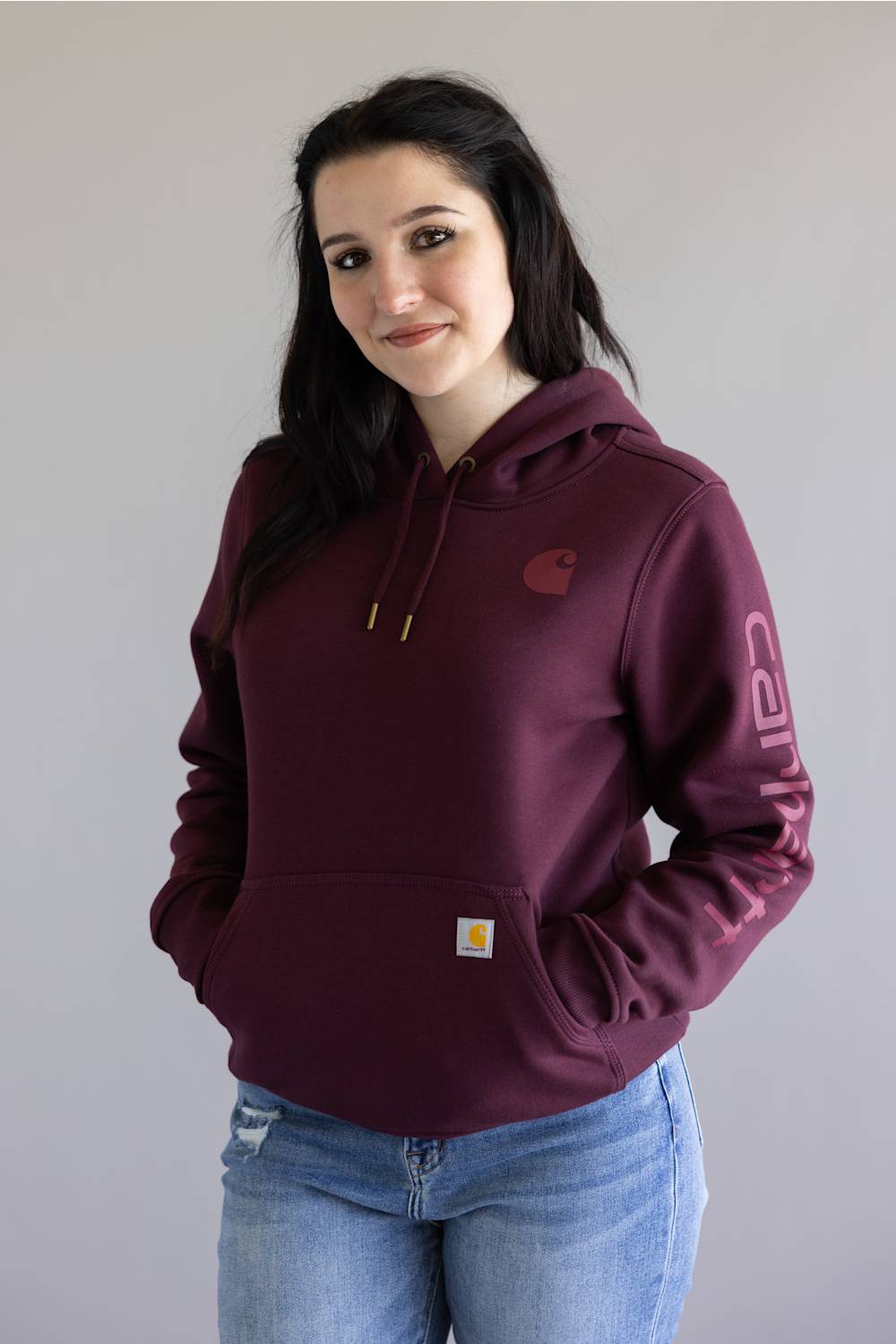 A woman with long dark hair smiling, wearing a deep cherrywood Carhartt hoodie with a front kangaroo pocket, a small chest logo, and the Carhartt logo printed along the left sleeve, paired with light-wash denim jeans.