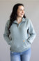 A woman wearing a light blue-gray quarter-zip sweatshirt with a small Carhartt logo patch on the front pocket, paired with blue jeans, standing against a neutral gray background.