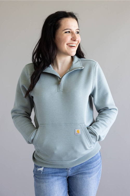 A woman wearing a light blue-gray quarter-zip sweatshirt with a small Carhartt logo patch on the front pocket, paired with blue jeans, standing against a neutral gray background.