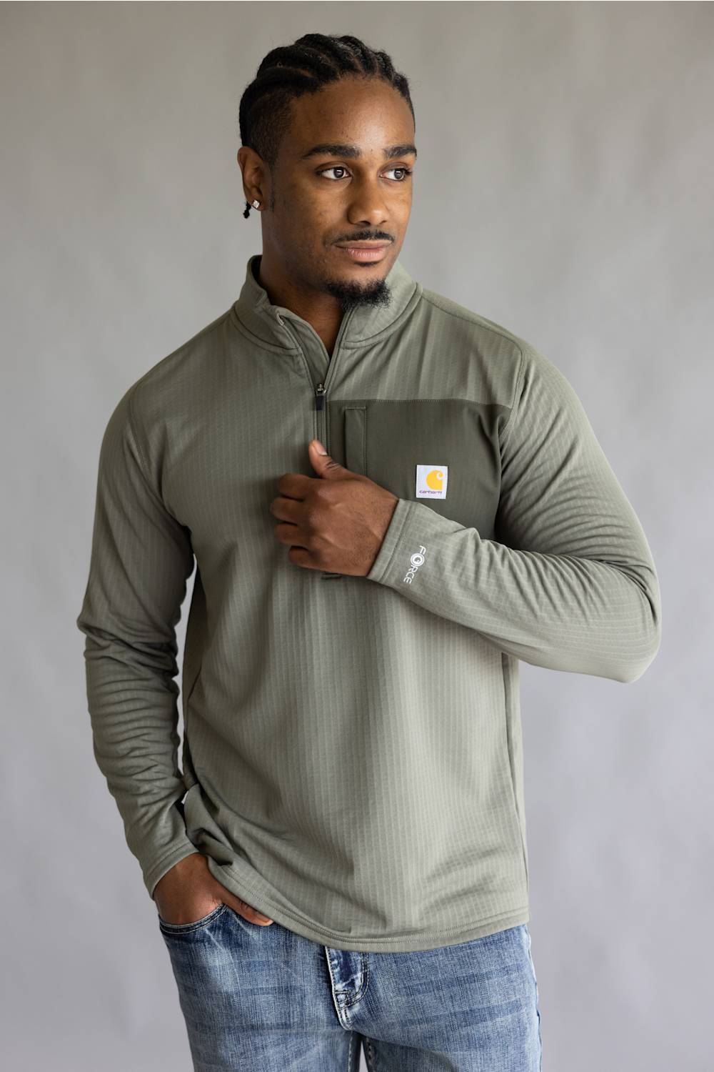 A man wearing a sage green ribbed long-sleeve quarter-zip pullover with a chest pocket and a small Carhartt logo patch, paired with blue jeans, against a neutral gray background.