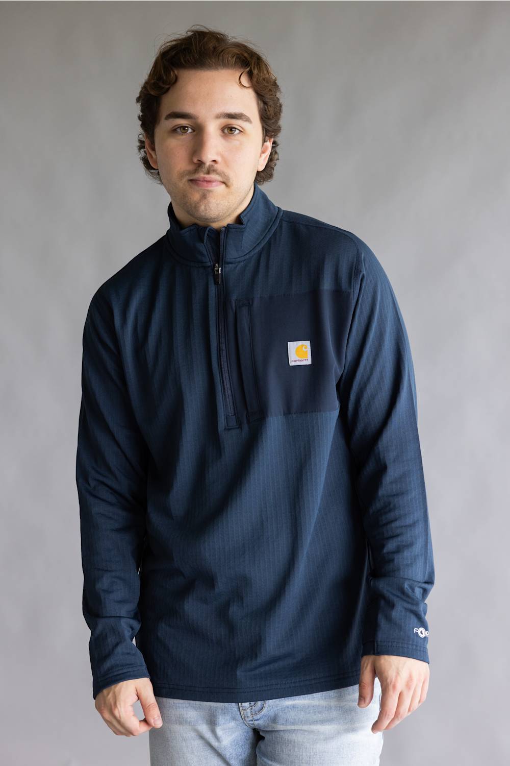 A man wearing a navy blue quarter-zip pullover sweatshirt with a small Carhartt logo on the left chest, paired with light blue jeans, standing against a neutral gray studio background.