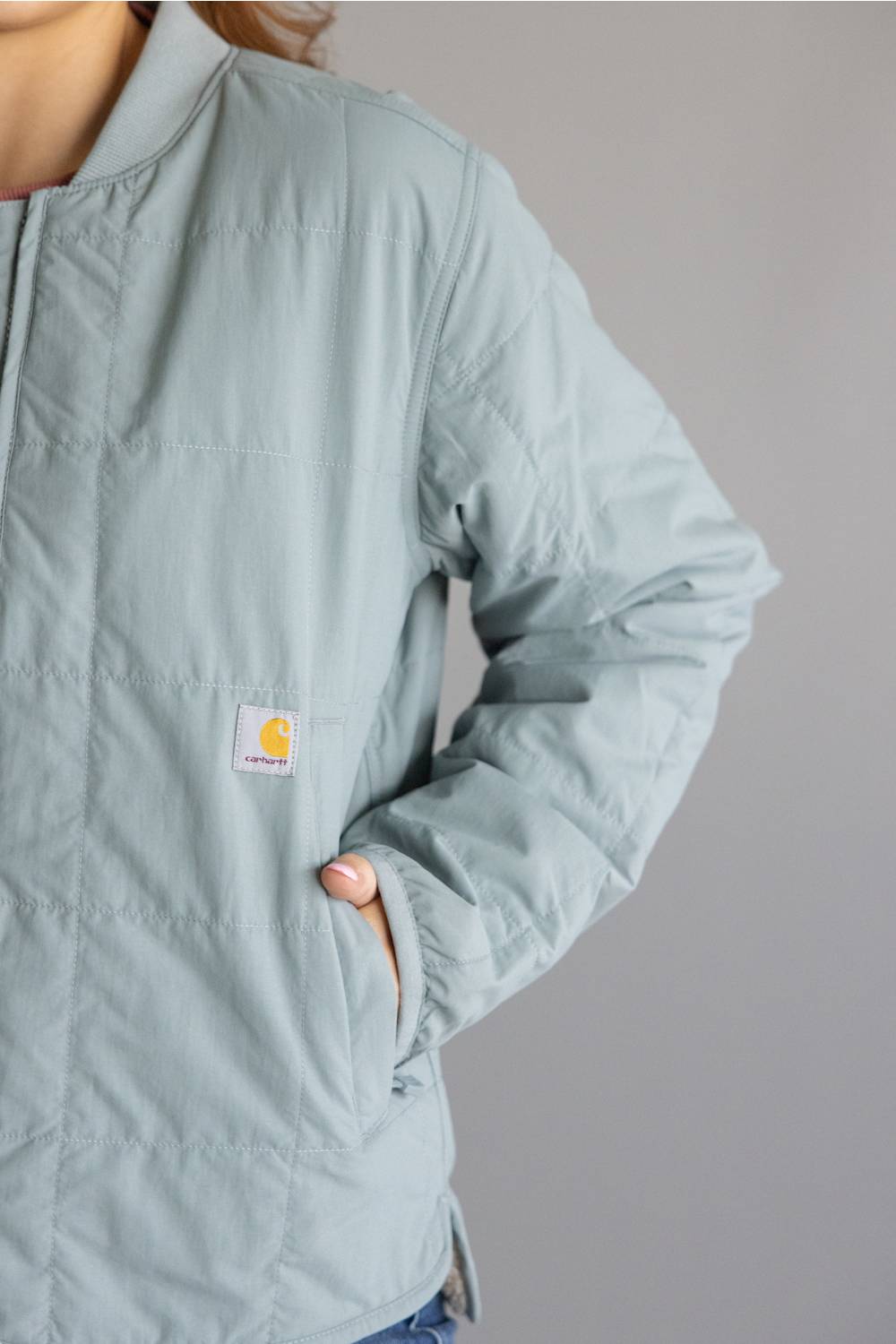A close-up of a person wearing a light sage green quilted jacket with a visible Carhartt logo patch on the chest, set against a plain gray background.