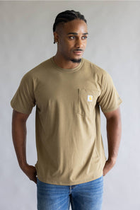 A man wearing a tan Carhartt short-sleeve crewneck pocket t-shirt and blue jeans, standing against a neutral gray studio background.
