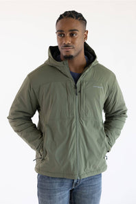A man wearing a dusty olive green hooded Carhartt jacket paired with blue jeans, posed against a plain white background with hands in his pockets.