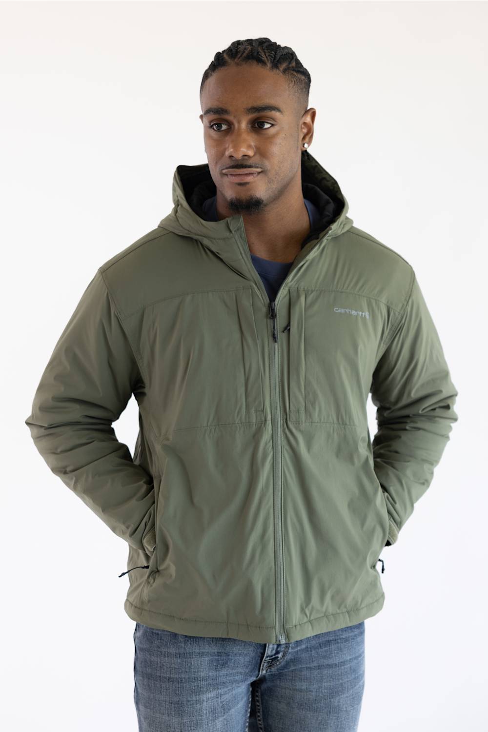 A man wearing a dusty olive green hooded Carhartt jacket paired with blue jeans, posed against a plain white background with hands in his pockets.
