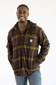 A smiling man wearing a brown hooded flannel shirt jacket with a gold plaid pattern, featuring a visible Carhartt logo patch on the chest, paired with blue jeans against a plain white background.