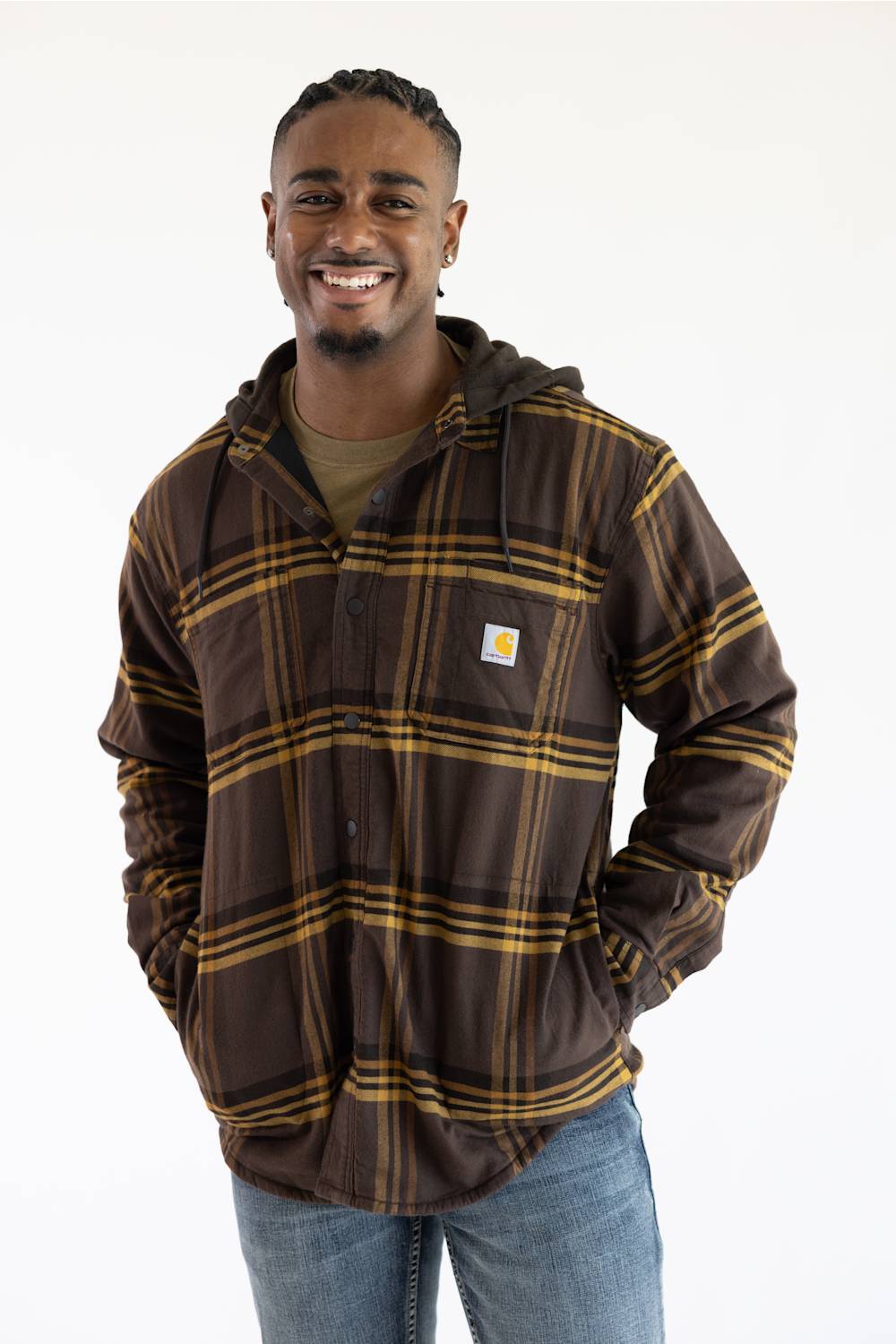 A smiling man wearing a brown hooded flannel shirt jacket with a gold plaid pattern, featuring a visible Carhartt logo patch on the chest, paired with blue jeans against a plain white background.