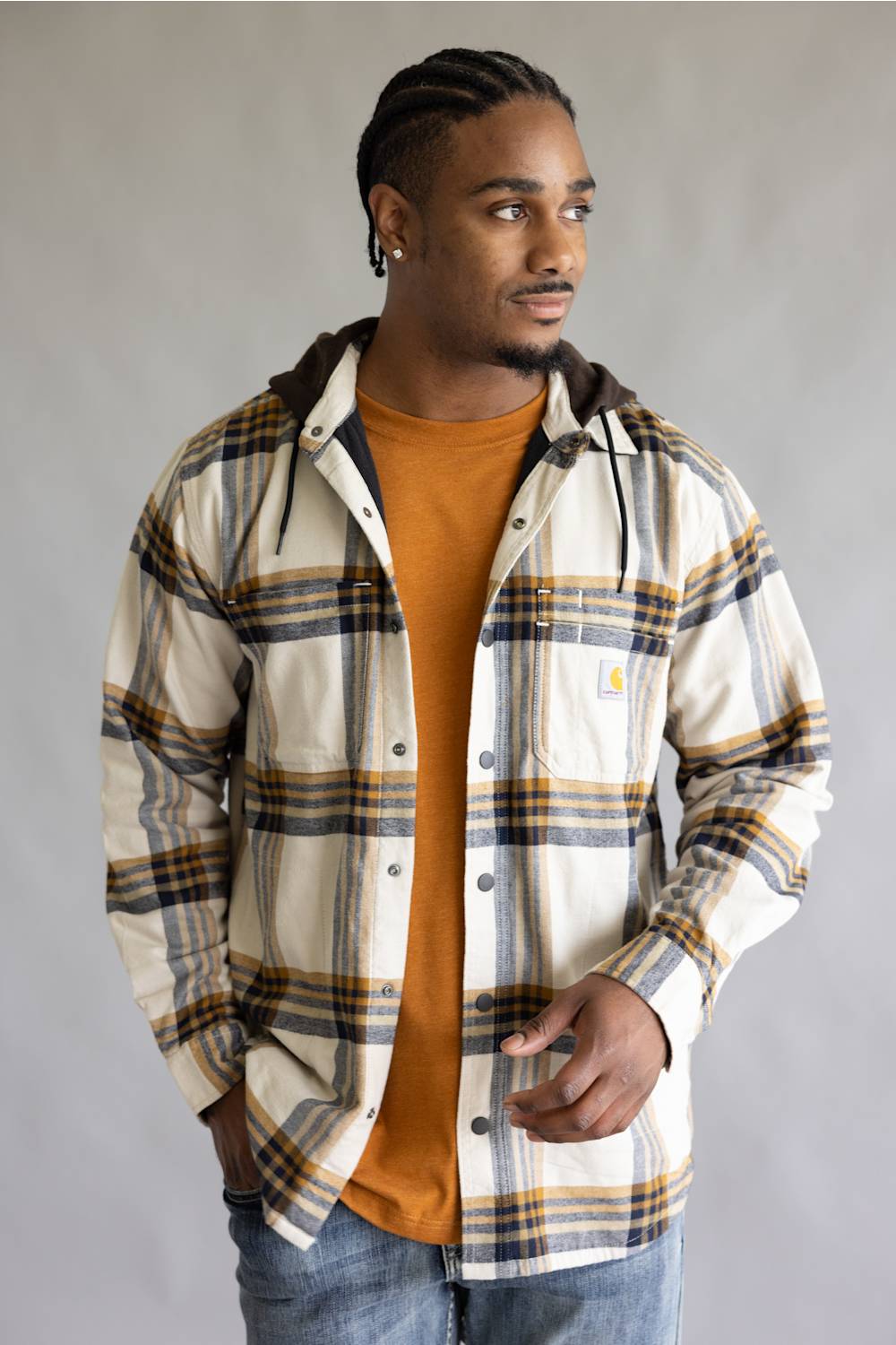 A man wearing a Carhartt hooded flannel shirt in a cream, navy, and mustard plaid pattern over an orange t-shirt and blue jeans, standing against a neutral gray background.