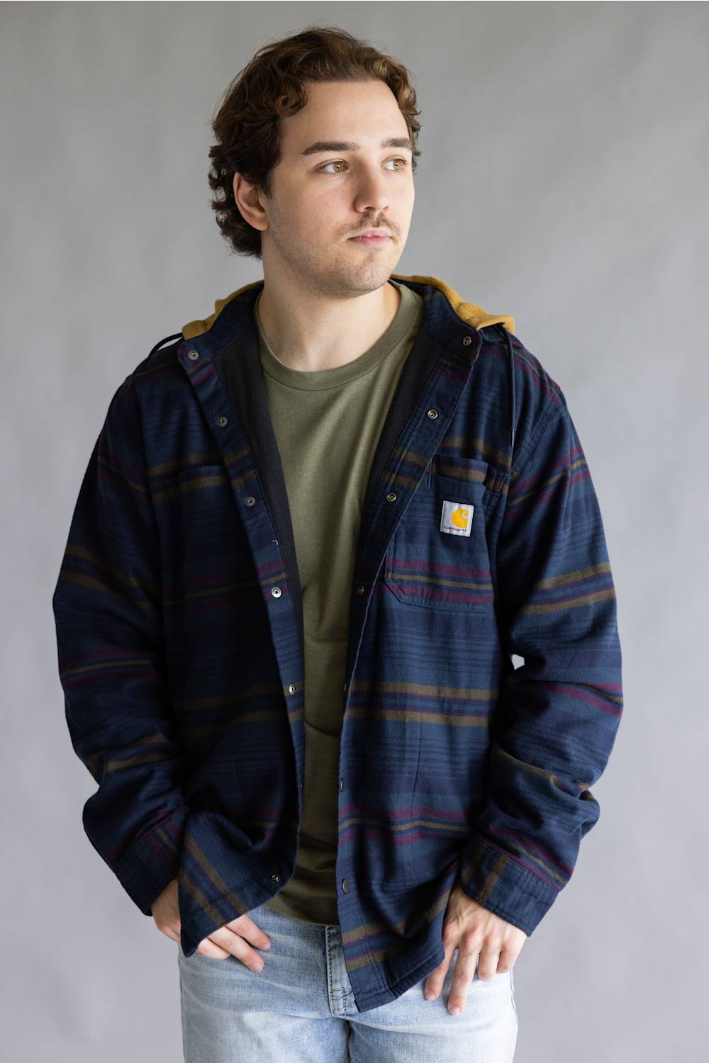 A man wearing a navy blue Carhartt plaid hooded jacket over an olive green t-shirt and light blue jeans, standing against a neutral gray studio background.