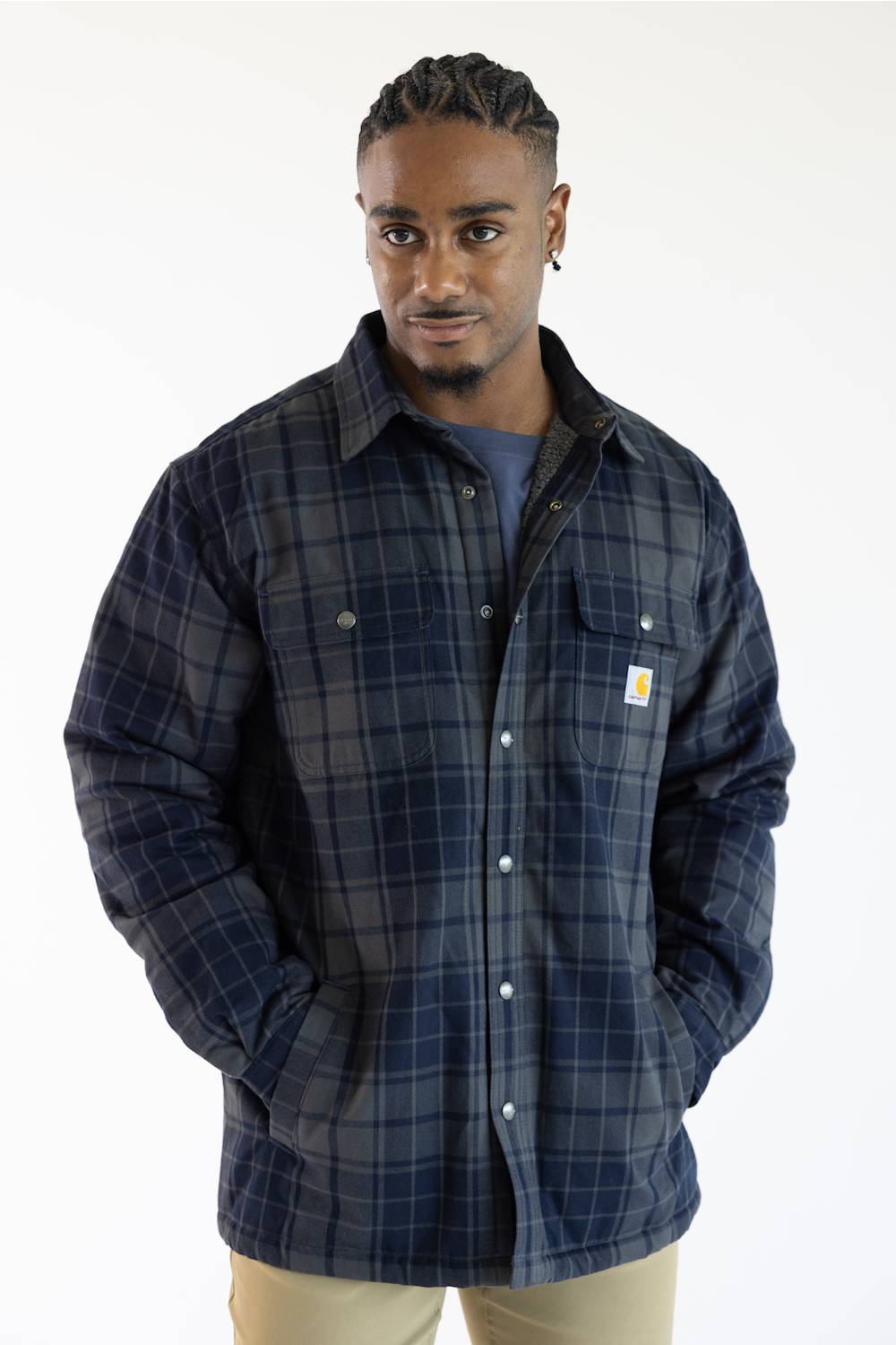 A man wearing a navy blue and gray plaid button-up overshirt with a small Carhartt logo patch on the chest pocket, paired with a blue t-shirt and beige pants, against a plain white background.