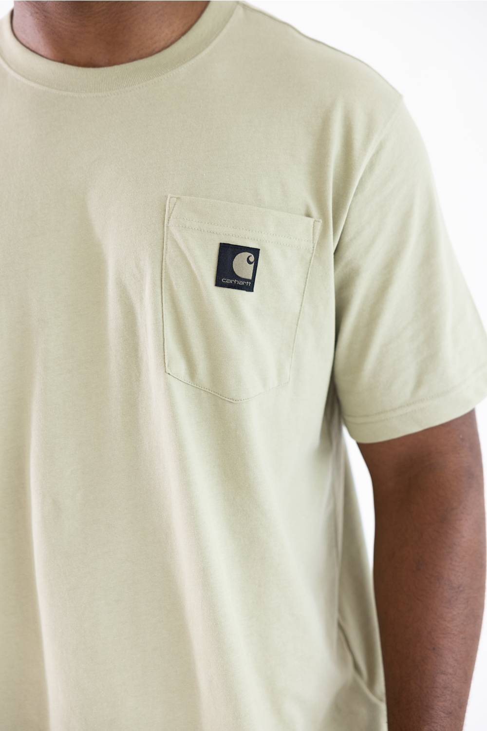A close-up of a person wearing a light cream-colored short-sleeve pocket t-shirt with a black Carhartt logo patch on the chest pocket.