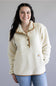 A smiling woman wearing a cream sherpa fleece pullover with tan contrast trim and a button placket, paired with blue jeans, against a plain gray studio background.