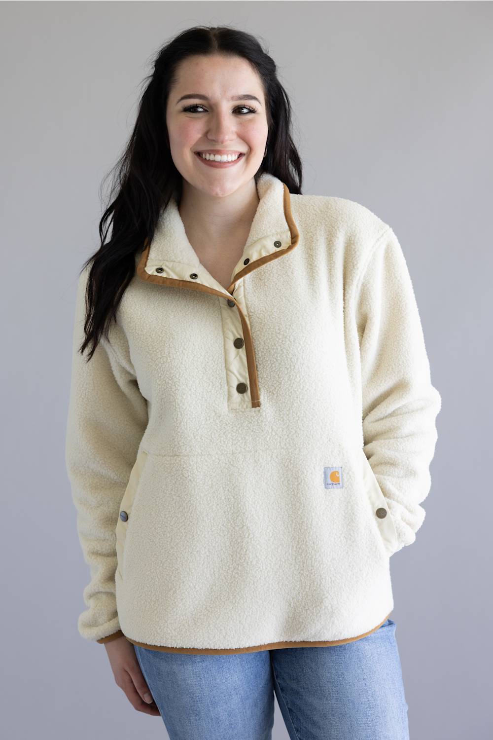 A smiling woman wearing a cream sherpa fleece pullover with tan contrast trim and a button placket, paired with blue jeans, against a plain gray studio background.