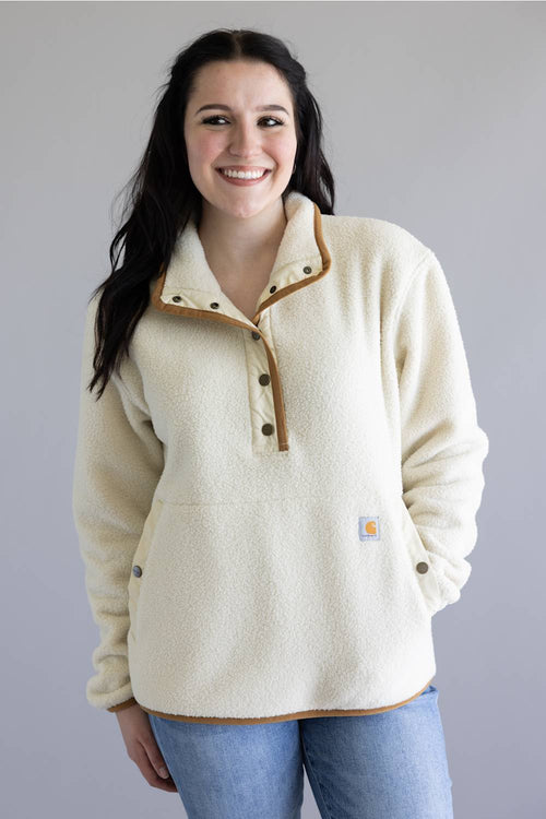 A smiling woman wearing a cream sherpa fleece pullover with tan contrast trim and a button placket, paired with blue jeans, against a plain gray studio background.