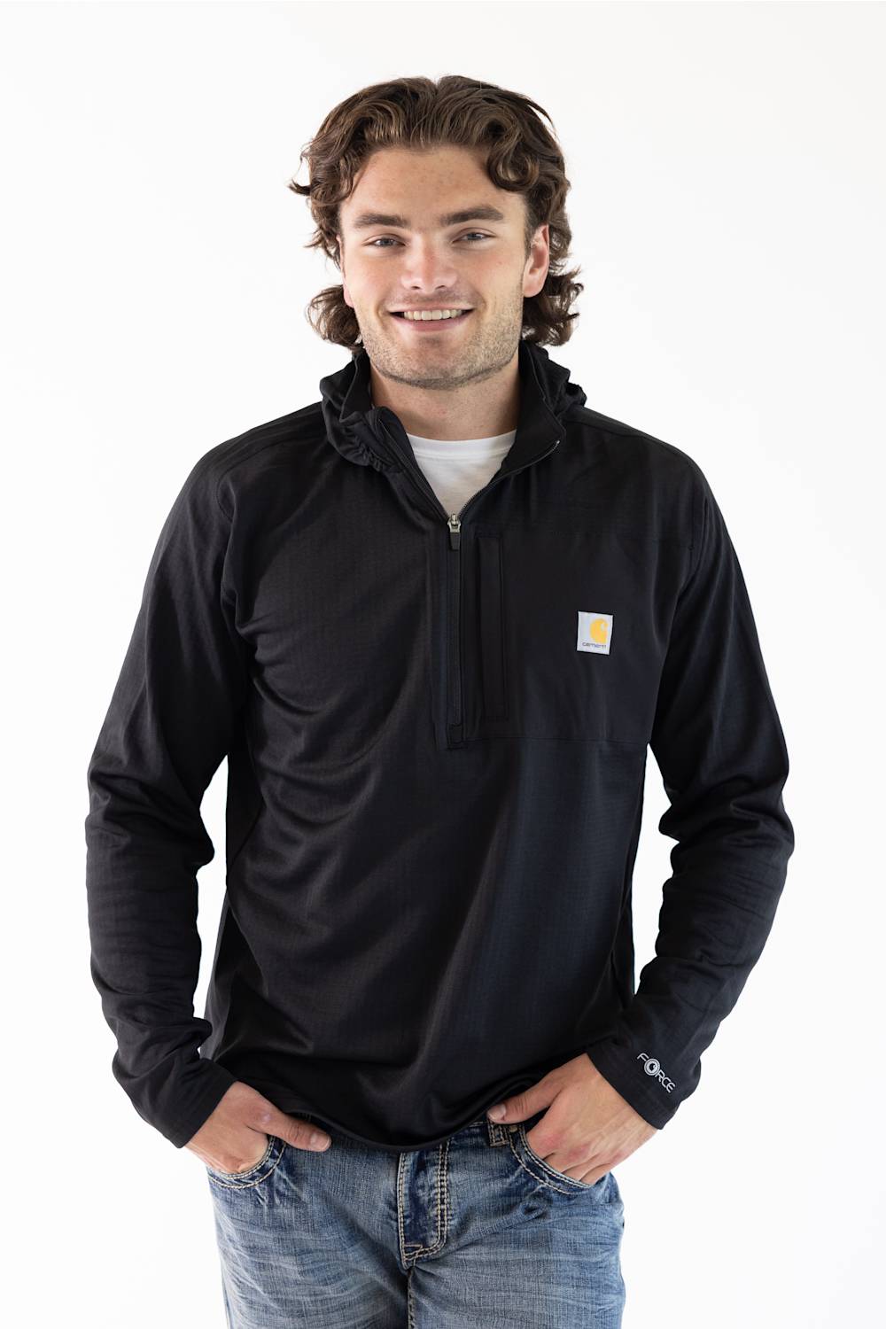 A male model standing against a white background, wearing a black Carhartt Force 1/2 zip hooded sweatshirt over a white t-shirt and blue jeans.