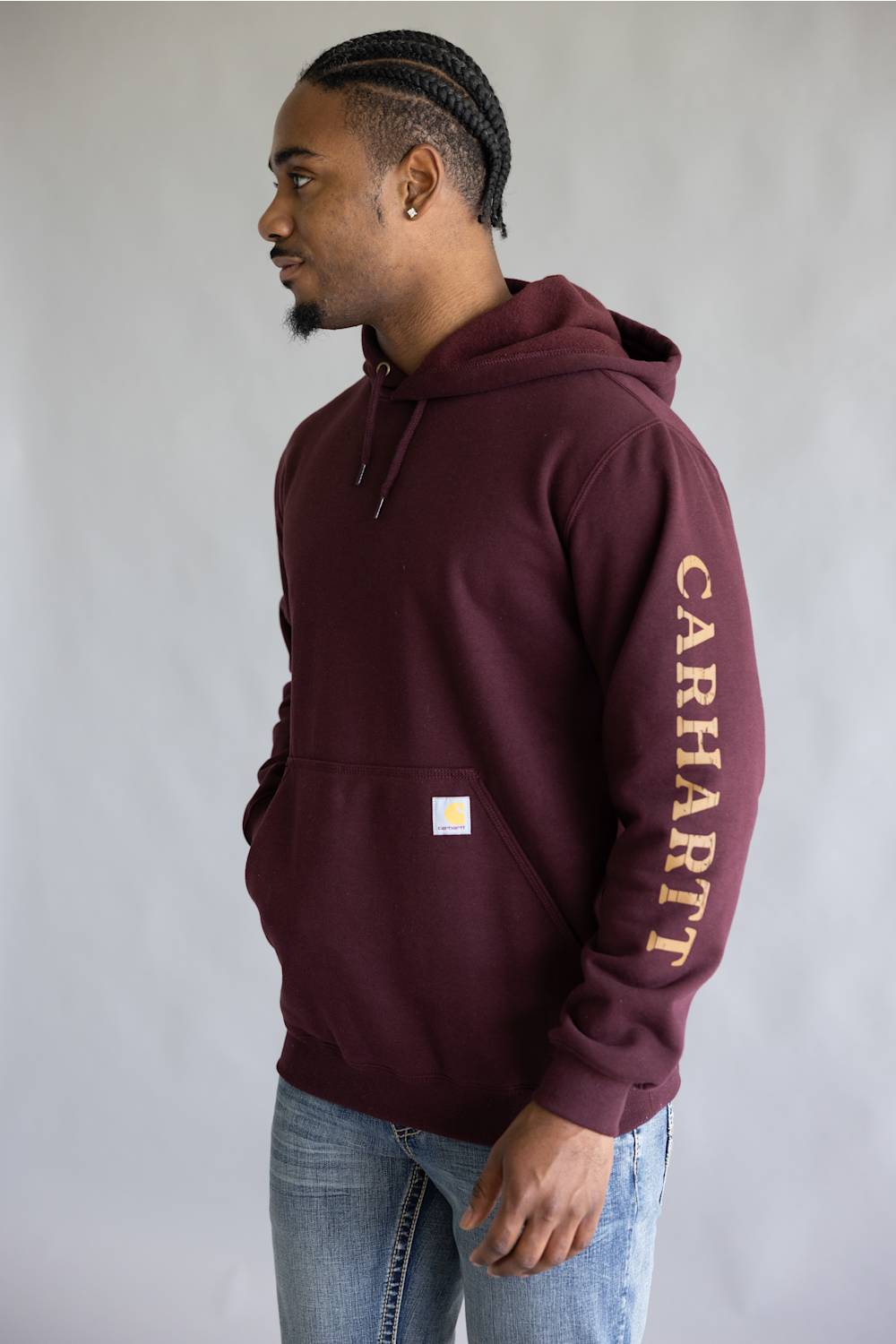 A man wearing a maroon Carhartt hoodie with gold lettering down the sleeve, paired with blue jeans, standing against a light gray background.