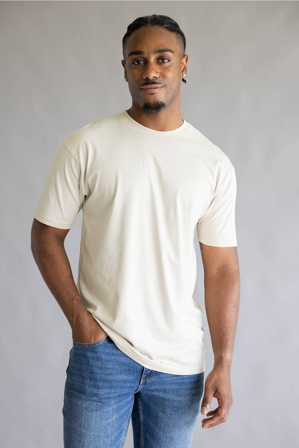 A man wearing a plain cream-colored short-sleeve crewneck t-shirt and blue jeans, standing against a neutral gray studio background.