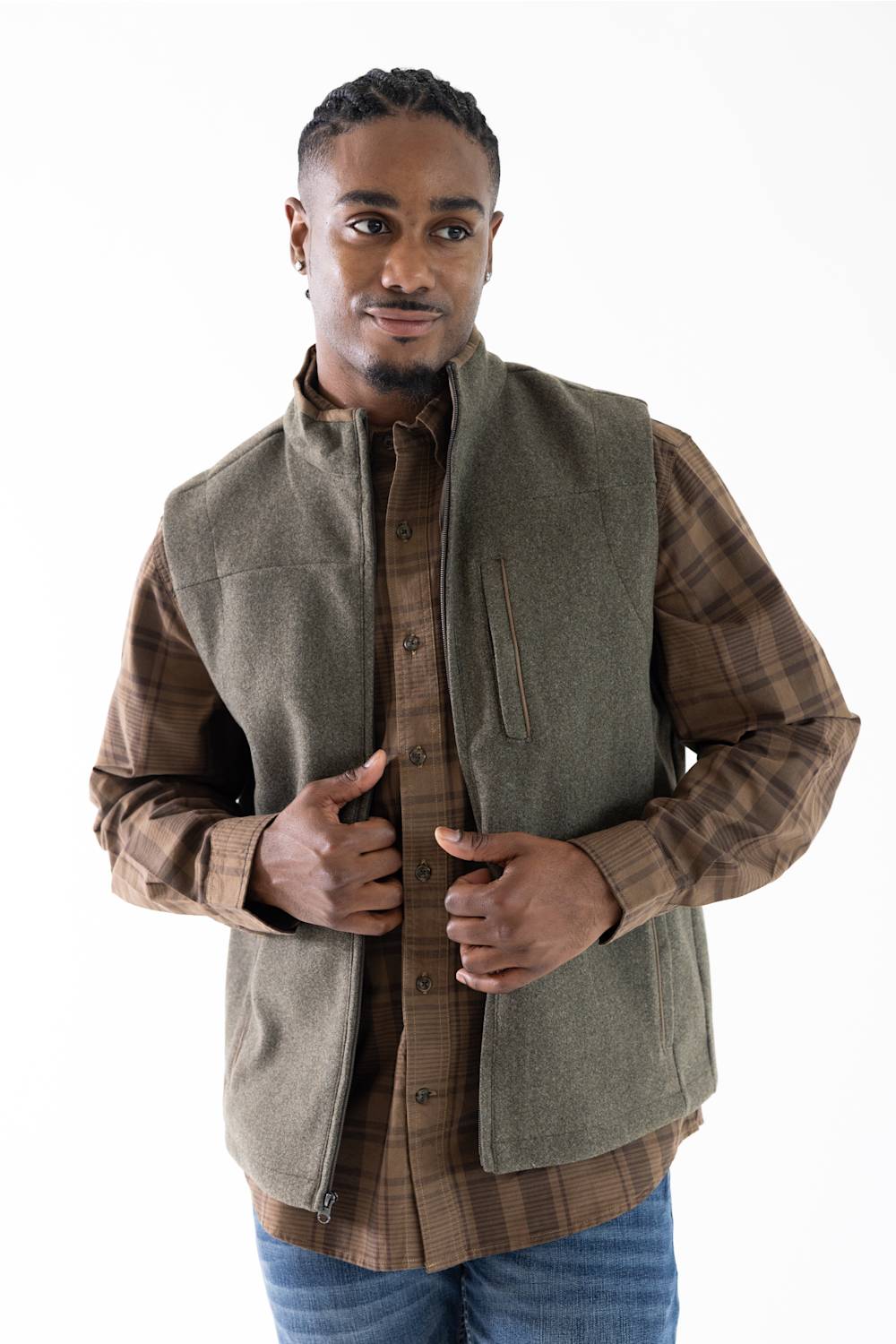 A man wearing a brown plaid button-down shirt with a olive green sleeveless vest layered over it, paired with blue jeans, against a plain white background.