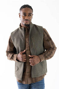 A man wearing a brown plaid button-down shirt with a olive green sleeveless vest layered over it, paired with blue jeans, against a plain white background.