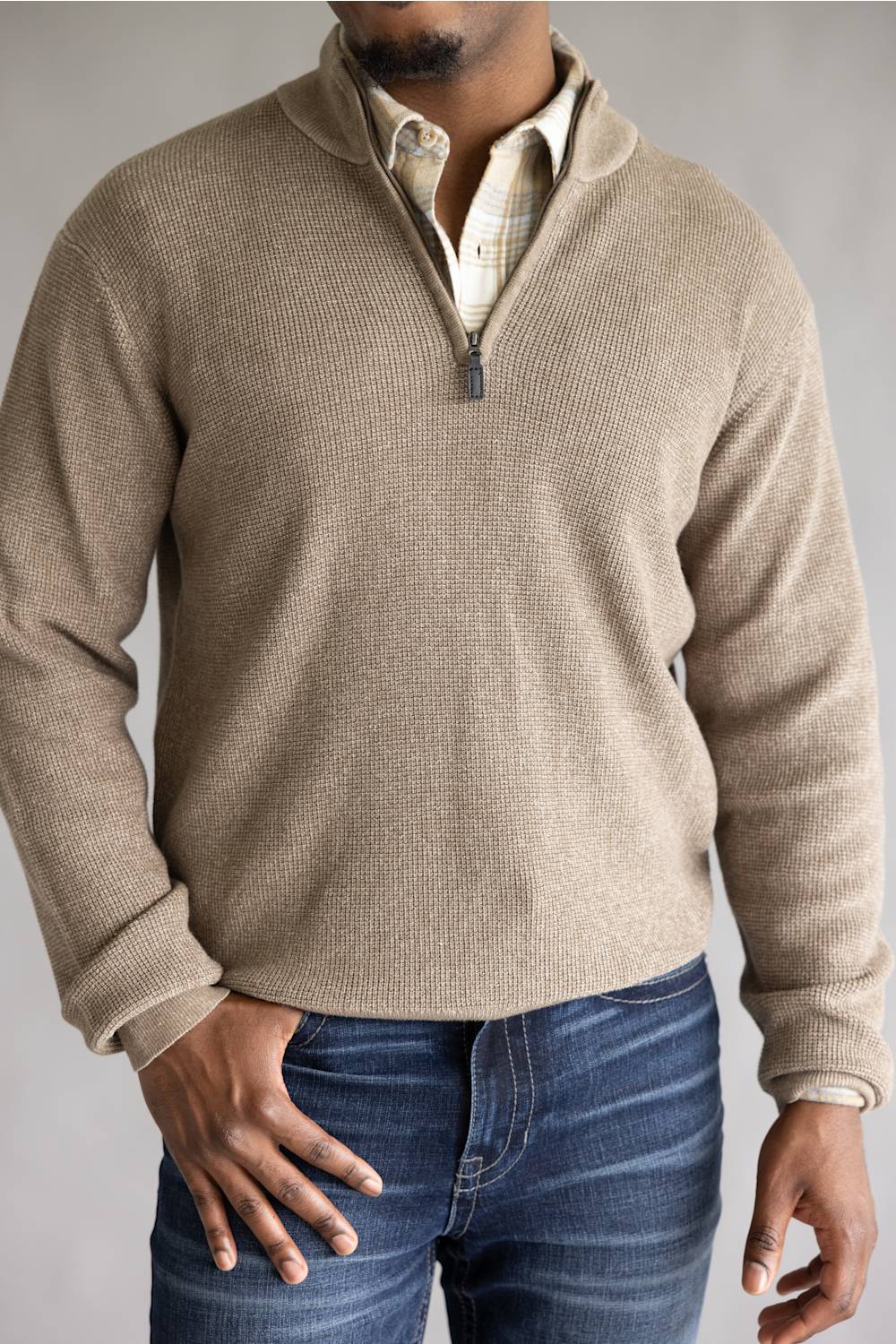 A man wearing a beige ribbed knit quarter-zip pullover sweater over a plaid shirt and blue jeans, shown from the neck down against a neutral gray background.