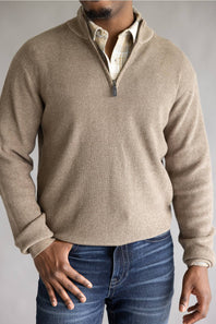 A man wearing a beige ribbed knit quarter-zip pullover sweater over a plaid shirt and blue jeans, shown from the neck down against a neutral gray background.