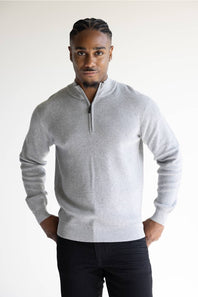 A man wearing a light gray ribbed knit quarter-zip pullover sweater and dark trousers, standing against a plain light background.