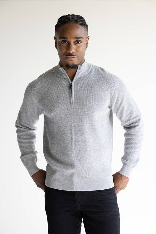 A man wearing a light gray ribbed knit quarter-zip pullover sweater and dark trousers, standing against a plain light background.