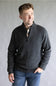 A man wearing a dark gray heathered quarter-zip pullover sweater over a plaid shirt and blue jeans, standing against a neutral gray studio background.
