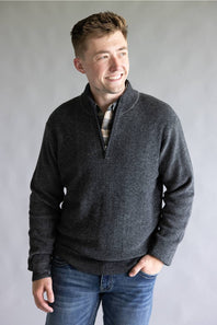 A man wearing a dark gray heathered quarter-zip pullover sweater over a plaid shirt and blue jeans, standing against a neutral gray studio background.