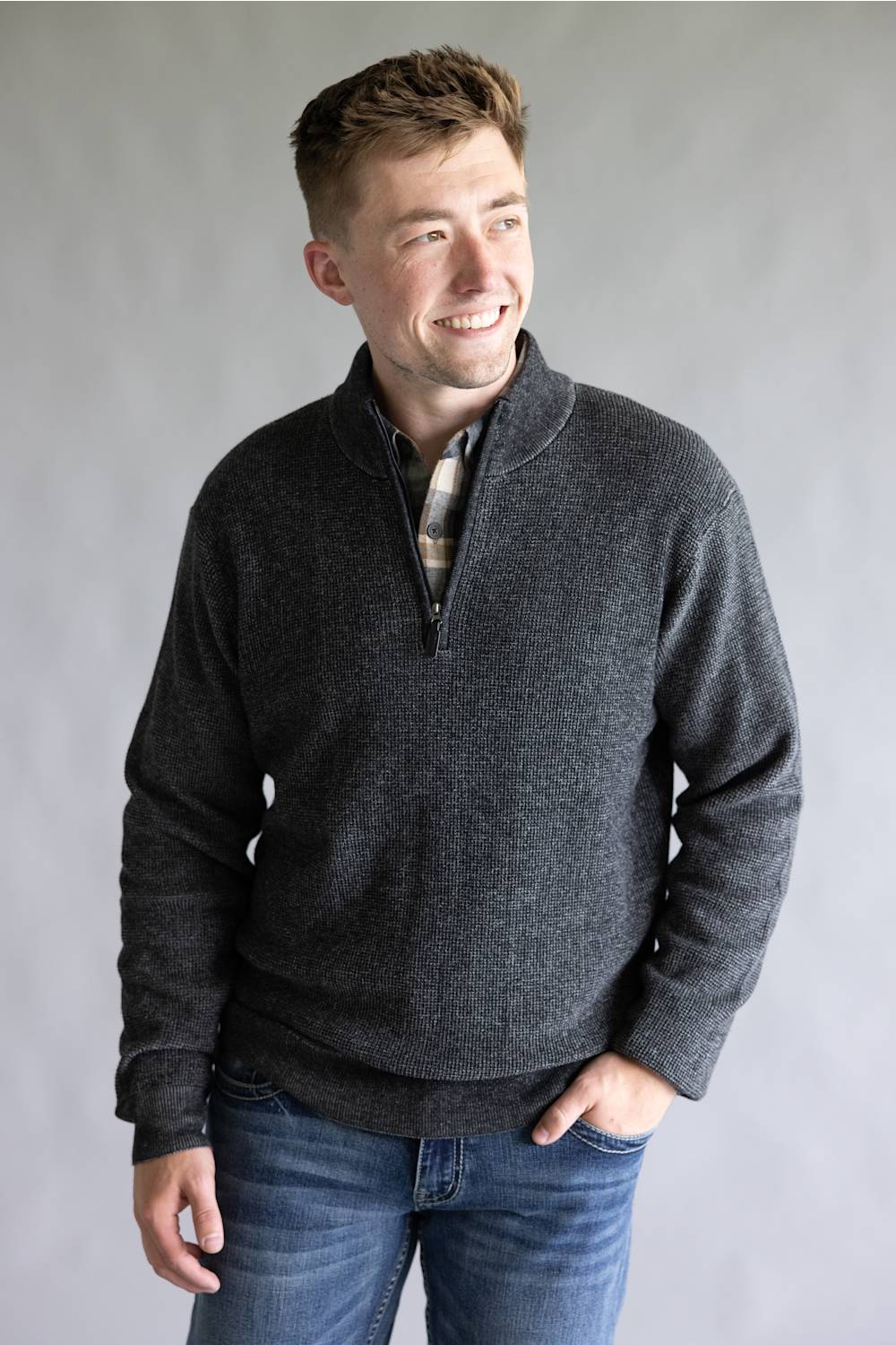 A man wearing a dark gray heathered quarter-zip pullover sweater over a plaid shirt and blue jeans, standing against a neutral gray studio background.