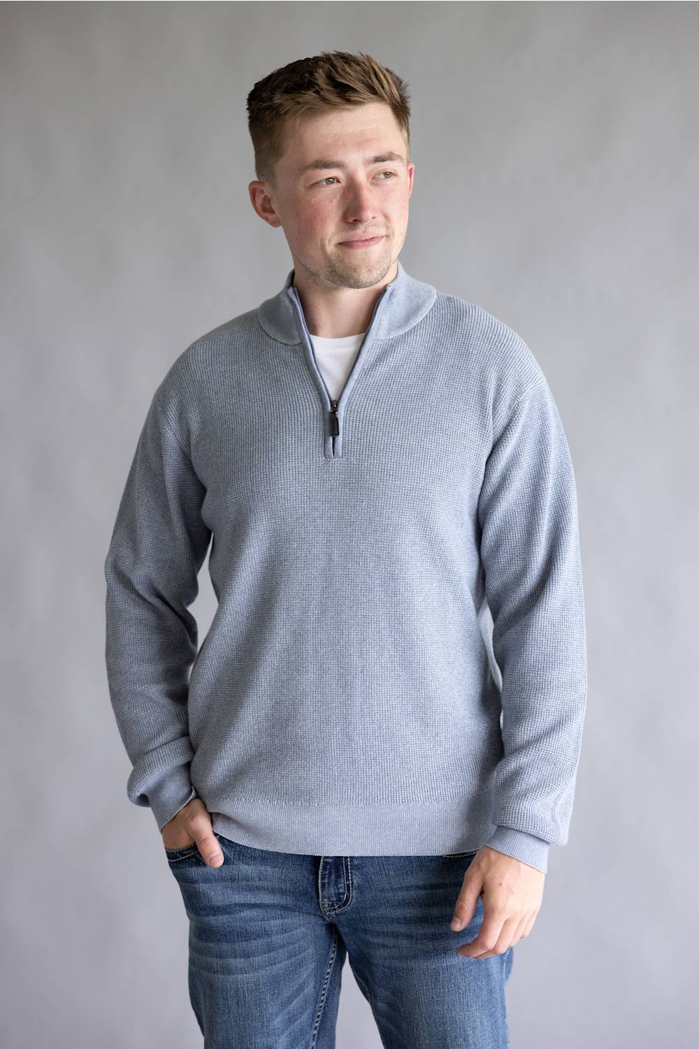 A man wearing a light gray quarter-zip pullover sweater over a white undershirt and blue jeans, standing against a neutral gray studio background.