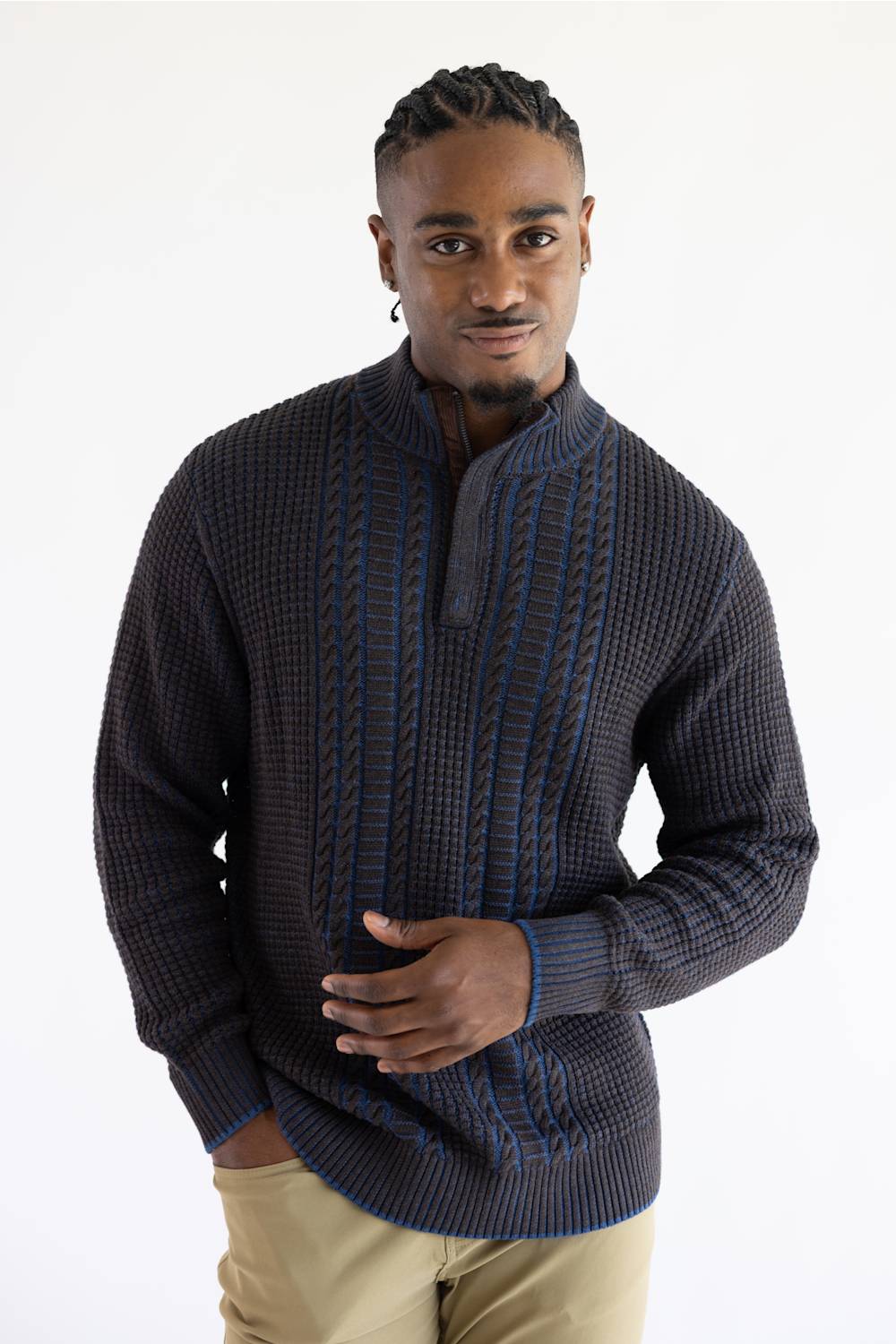 A man wearing a dark charcoal gray and navy blue textured knit sweater with a mock neck and blue cable-knit accents, paired with khaki pants, against a plain white studio background.