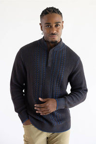 A man wearing a dark charcoal gray and navy blue textured knit sweater with a mock neck and blue cable-knit accents, paired with khaki pants, against a plain white studio background.
