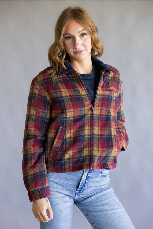 A woman wearing a red, yellow, and blue plaid flannel jacket over a dark blue shirt and light blue jeans, standing against a neutral gray studio background.