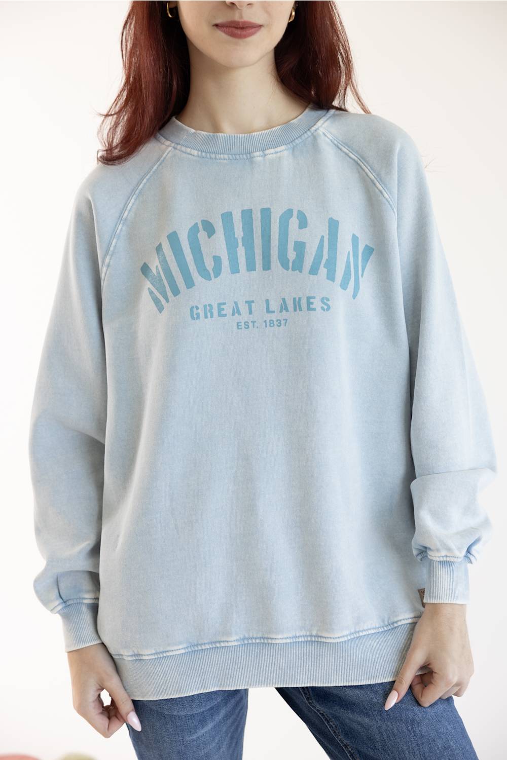 A woman wearing a light blue washed sweatshirt with 'MICHIGAN GREAT LAKES EST. 1897' printed in blue on the front, paired with blue jeans against a plain white background.