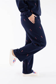 Side view of a woman wearing navy blue straight-leg fleece sweatpants featuring an all-over embroidered red and white candy cane pattern, with white sneakers and a matching sweatshirt.