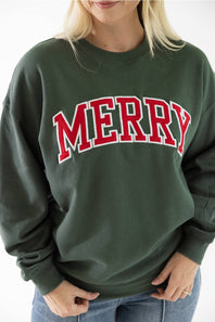 Detail shot of a woman wearing a dark green crewneck fleece sweatshirt featuring red and white block lettering that reads "Merry" across the chest.