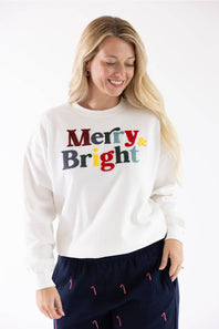 Studio portrait of a blonde woman wearing an ivory crewneck sweatshirt with colorful chenille "Merry & Bright" lettering and dark blue holiday-patterned lounge pants.