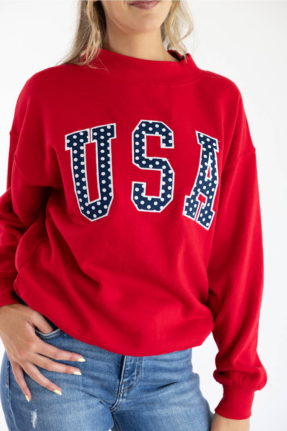 Close-up front view of a woman wearing the red 1897 Active USA mock neck sweatshirt, featuring navy blue "USA" applique lettering filled with white polka dots across the chest.