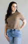A woman modeling a short-sleeve crewneck knit top in a warm taupe-brown color, paired with light-wash blue jeans, against a solid light gray background.