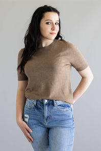 A woman modeling a short-sleeve crewneck knit top in a warm taupe-brown color, paired with light-wash blue jeans, against a solid light gray background.
