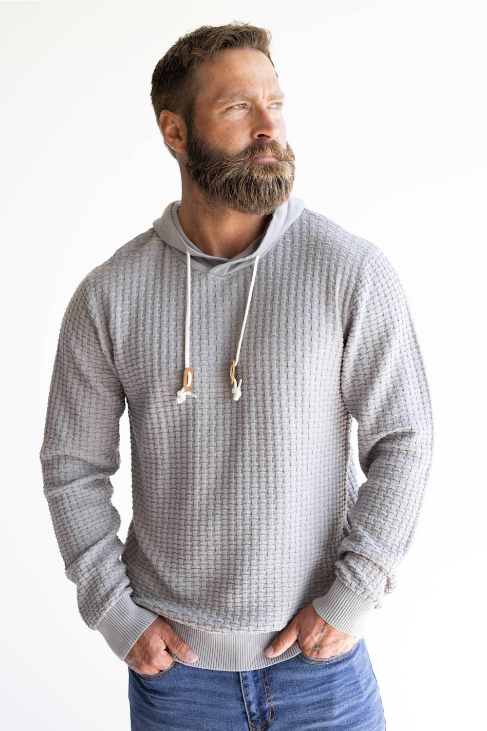A man with a full beard wearing a light gray waffle-knit hoodie with white drawstrings, paired with blue jeans, standing against a plain white background.
