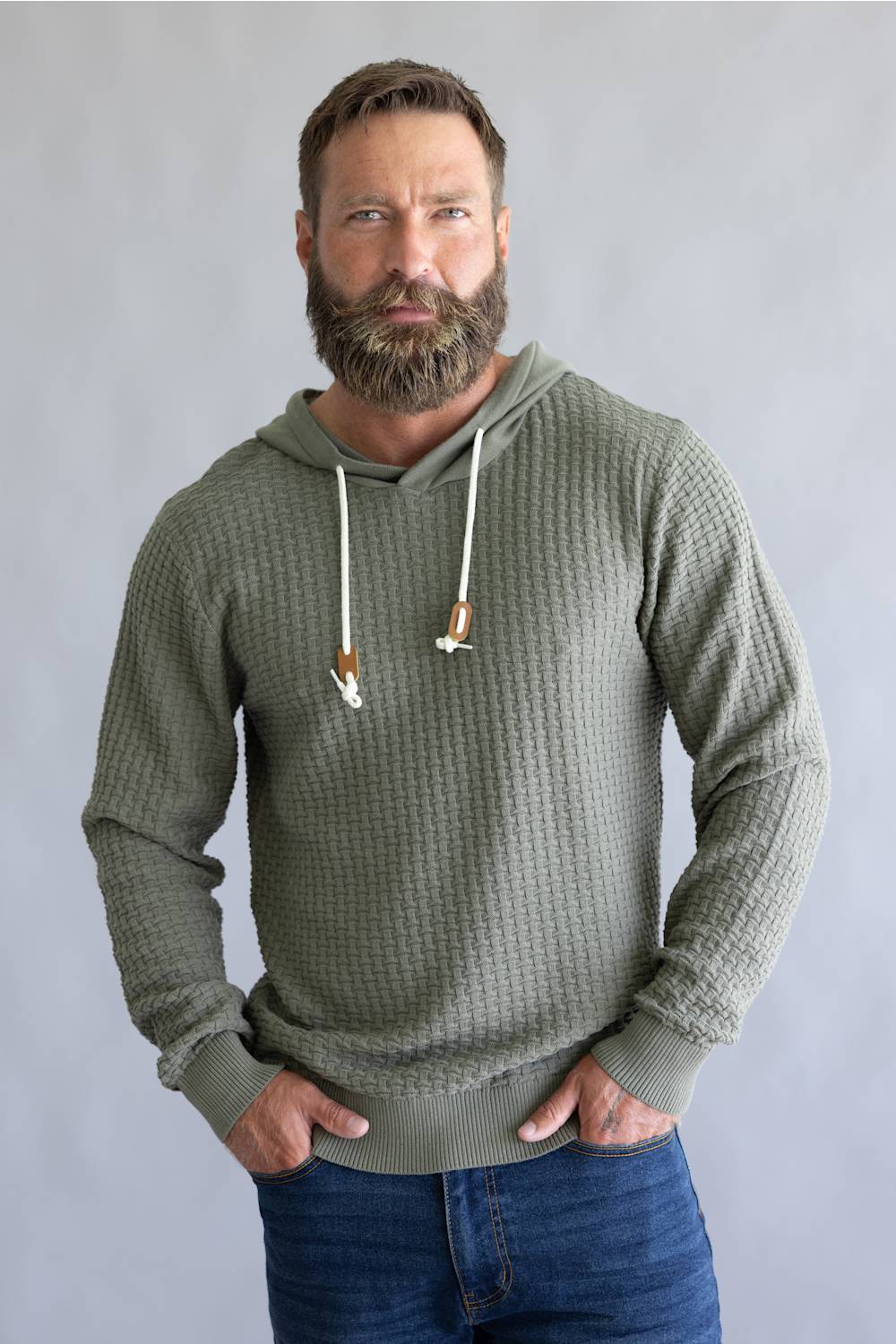 A man with a full beard wearing an olive green waffle-knit hoodie with white drawstrings, paired with blue jeans, standing against a neutral gray background.