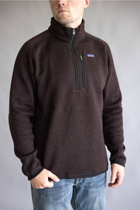 A man wearing a dark brown Patagonia quarter-zip fleece pullover sweater with blue jeans, standing against a neutral gray studio background.