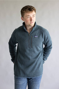 A man wearing a heathered slate blue quarter-zip fleece pullover with the Patagonia logo on the chest, paired with blue jeans, standing against a neutral gray background.